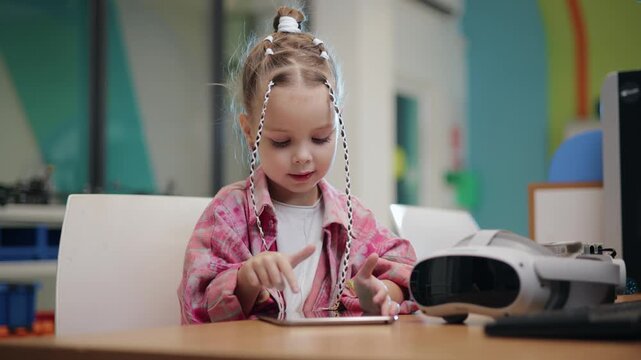 Slow motion preschool girl tapping tablet at colorful classroom learning table with vr headset beside her.