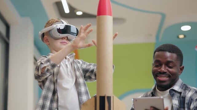 Kid boy testing space in vr headset while adjusting model rocket in makerspace with mentor teacher using tablet.