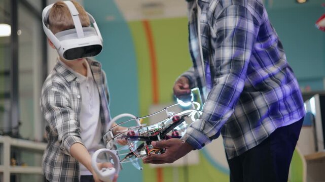 Kid boy interacting with drone and vr headset in makerspace workshop while teacher man assists handheld follow.