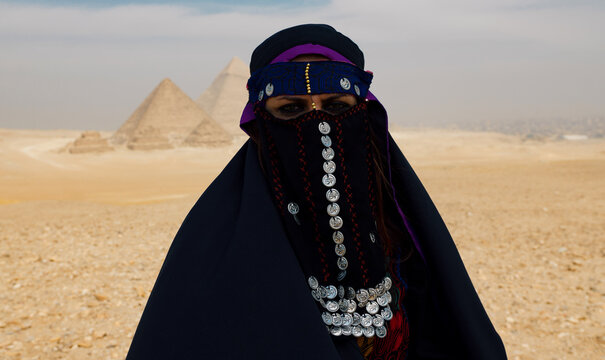 Striking Bedouin woman in traditional niqab and silver coin veil posing before the Great Pyramids of Giza in the Egyptian desert ideal for travel