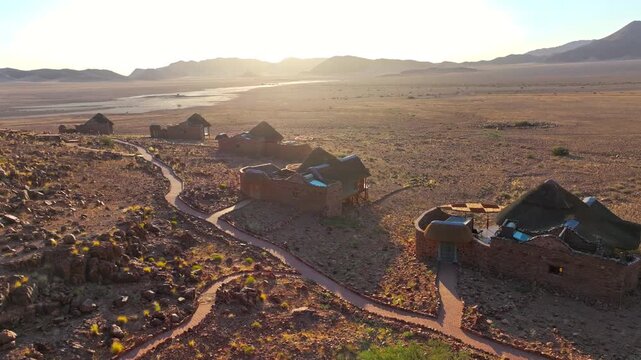 Drone view of a safari lodge of stone and thatch buildings spread across the red Namib desert. The remote eco retreat blends into the red desert landscape beneath wide skies and distant mountains.