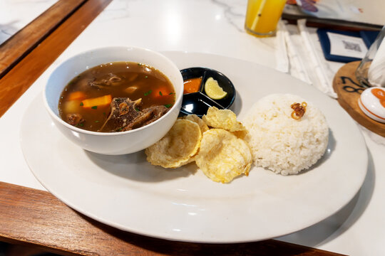 Indonesian oxtail soup or Sop Buntut served with a bowl of white rice, emping crackers, chili sauce, and lime lime on a white plate.