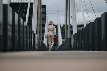 Fototapeta premium Businesswoman walking away across a modern bridge carrying a red bag. A lone professional crossing an urban walkway in a neutral, overcast setting.