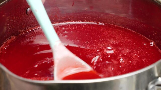 Red currant jam boils and bubbles intensely in a hot stainless steel pot. Sugar and fresh juice are mixed with a spatula. Thick red confit simmers during the traditional cooking process.
