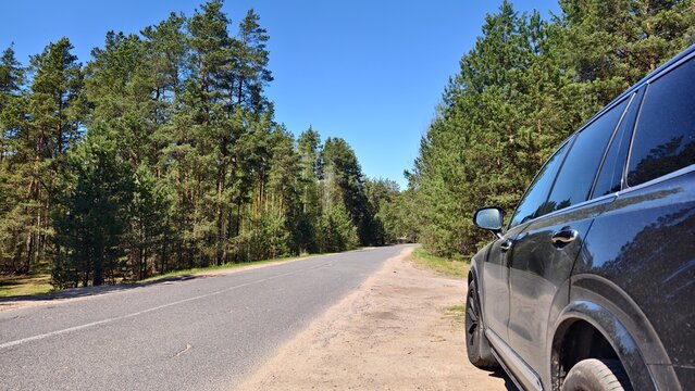 A short stop during a spring trip. The car is parked on the sandy roadside of an asphalt road leading through the forest. Nearby there are pines, birches with young foliage and a turn in the road. Sun