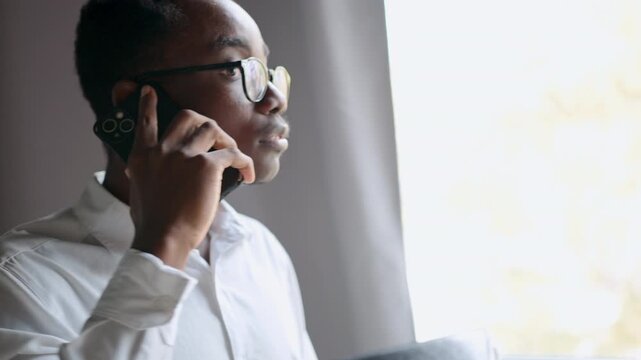 A black man in glasses expresses his thoughts on errors made by his team during a phone call. He engages in a serious conversation while working in an office, focusing on resolving issues