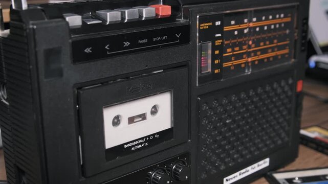 Close-up of a finger pressing play and stop buttons on a black retro tape recorder with white spinning reels. German text and an orange radio tuner dial are visible in the background.