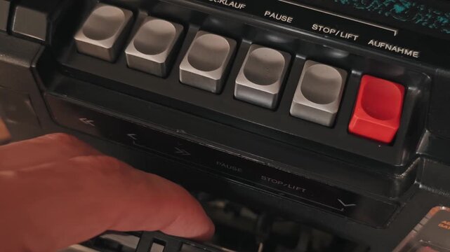 Close-up of a finger pressing the silver stop lift button on a retro tape deck to eject a cassette. The German text controls and textured black panel highlight the mechanical quality of the device.