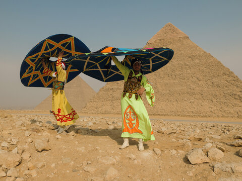 Two traditional folk dancers spin large colorful hats in vivid costumes before the Giza pyramids in Egypt celebrating cultural performance and tourism