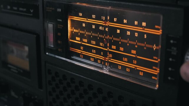 Extreme close-up of a vintage radio tuner dial with a red indicator moving across an orange frequency scale. The search for a station represents the precision of analog broadcast technology.