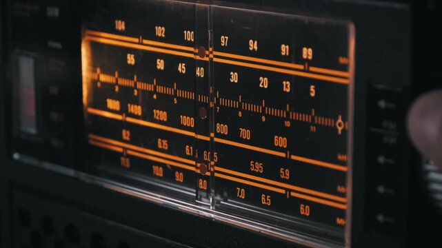 Side view of a hand turning the tuning knob on an old radio receiver to search for a station. The indicator moves across the illuminated orange frequency scale to highlight classic analog technology.