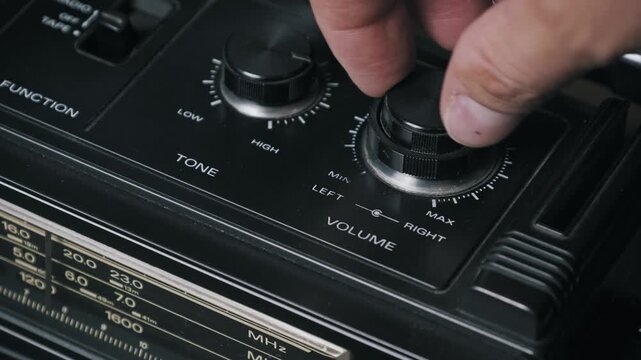 A male hand rotates the volume dial clockwise on a black vintage radio to increase the sound. Concept of nostalgia, broadcasting, and adjusting audio settings on old technology.