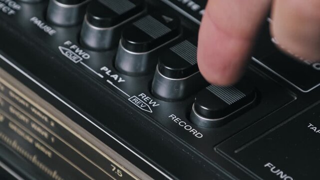 Angled macro shot of a finger pressing a record button on a vintage cassette player. The black plastic controls and silver accents highlight the retro boombox design.