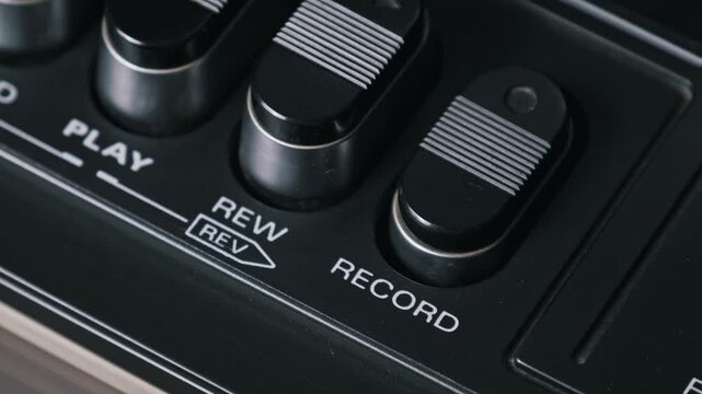 Extreme close-up top-down view of a finger pressing the record button on an old tape recorder. The textured black keys and silver accents highlight the vintage design.
