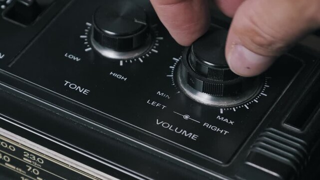 Close-up of a hand adjusting a black volume knob on a vintage radio cassette player. The dusty control panel and tone knob highlight the retro audio equipment.