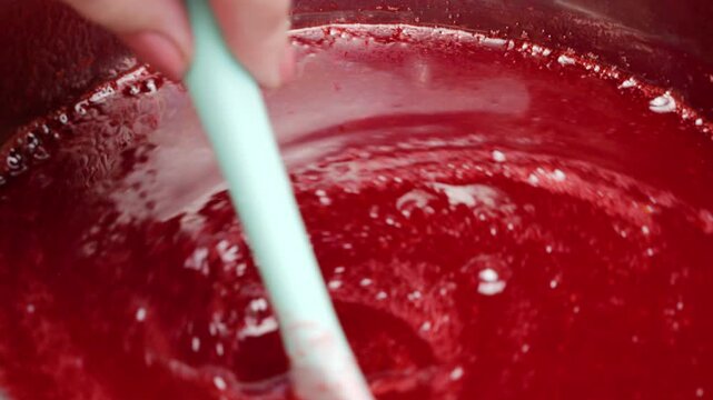 Red currant jam boils and bubbles intensely in a hot stainless steel pot. Sugar and fresh juice are mixed with a spatula. Thick red confit simmers during the traditional cooking process.