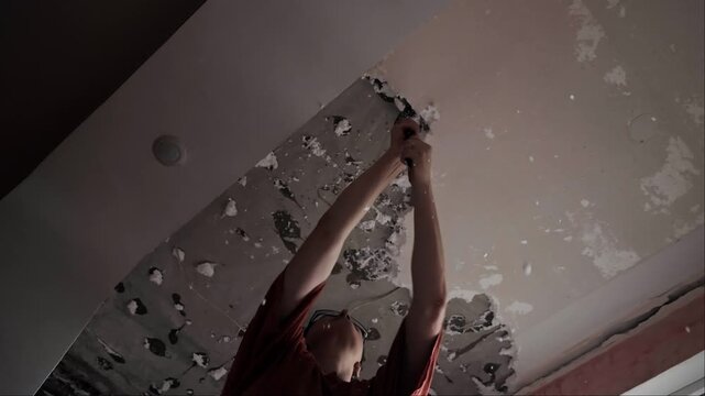 Low angle view of woman removing ceiling finish, chipping away surface coating during interior renovation and home repair work process.