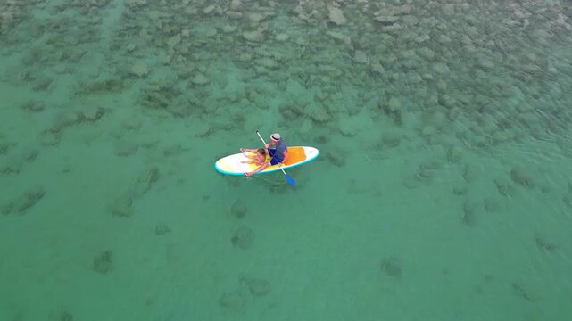 High angle drone shot of a loving father and his little daughter paddling together on a stand up paddleboard in clear turquoise sea water over a rocky bottom. High quality 4k footage