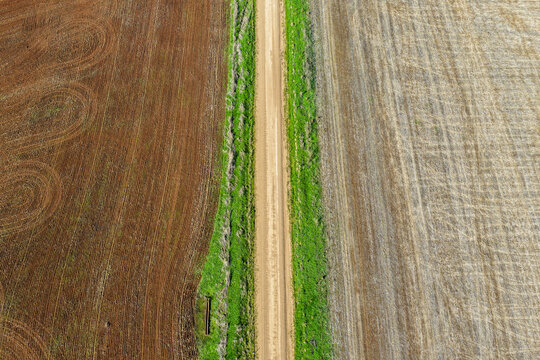 Aerial downward view of a green grass lined dirt road leading through cultivated agricultural fields, showcasing rural farmland patterns and textures beneath soft natural light in NSW, Australia.