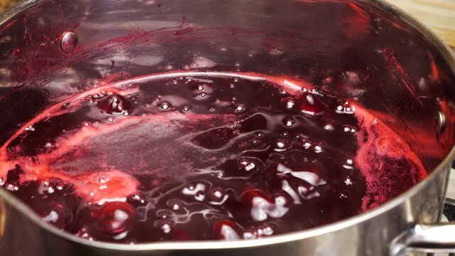 Boiling red currant jam bubbles in a saucepan during the home preparation process. Fresh berry confit cooks on the stove. Thick red jelly with small bubbles is simmering into sweet preserve.