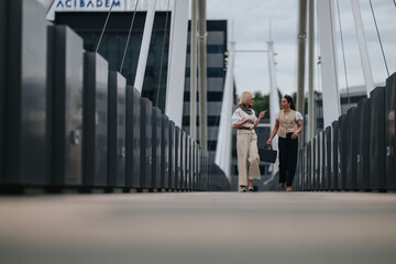 Fototapeta premium Two businesswomen walk and talk across a modern pedestrian bridge carrying a briefcase. The colleagues converse while crossing an urban suspension bridge in professional attire.