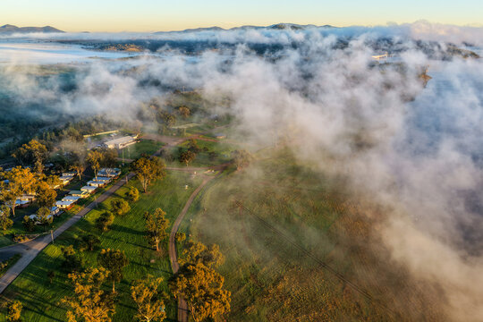 Aerial view over the Lake Keepit campgrounds as fog drifts across the surrounding rural landscape beneath soft sunrise light in NSW, Australia.