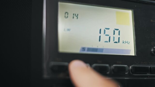 Close-up of a finger rapidly switching between MW and SW radio bands on a digital receiver. The LCD screen shows changing frequency units before settling on the medium wave band.