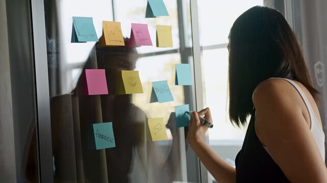Indian woman writes ideas on colorful sticky notes stuck to a glass wall in an office during a brainstorming session. She focuses on developing new concepts during the daytime.