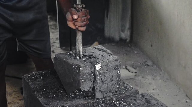 A person manually chisels a heavy, black block, possibly coal or briquette, creating dust and debris in an industrial setting.