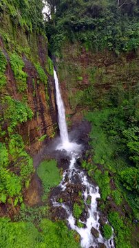 Cinematic FPV drone flight toward Kapas Biru Waterfall through lush tropical rainforest in East Java, Indonesia. Dramatic jungle landscape with a tall cascading waterfall and mist.