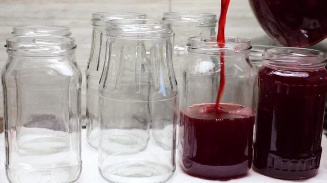Red currant jam is poured from a glass pitcher into sterile jars during home canning. Thick dark red syrup flows from a transparent jug. Sweet berry confit prepares for winter storage.