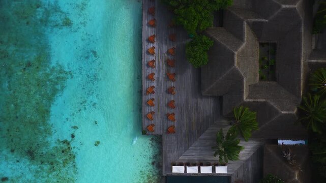 Aerial view of a Maldives beachfront resort with a long wooden deck, orange lounge chairs, thatched pavilions, coconut palms, and a small pool by a turquoise lagoon.