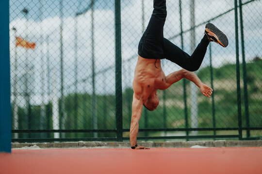 Shirtless athletic man balancing in a one-arm handstand on a fenced outdoor court. The male athlete practices strength and balance in an urban sports setting.