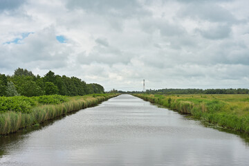 Fototapeta premium Straight Dutch canal flanked by grassy banks and trees under an expansive cloudy sky.