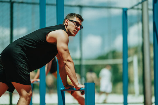 A fit young man leans on parallel bars performing an outdoor workout. The athlete trains strength and balance during a sport exercise session.