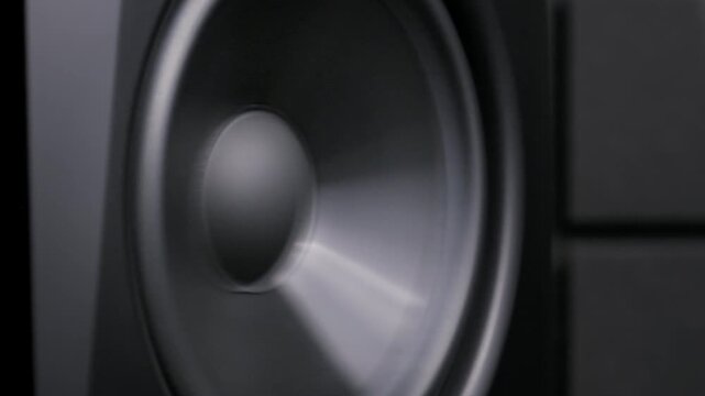 Close-up of a black speaker cone vibrating slowly while generating powerful sound waves. The dynamic driver creates rhythmic low-frequency pulses in a music studio.