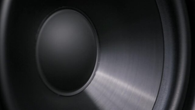 Macro perspective of a black subwoofer diaphragm vibrating intensely from low-frequency bass. The close-up reveals membrane texture and dynamic motion in a studio.