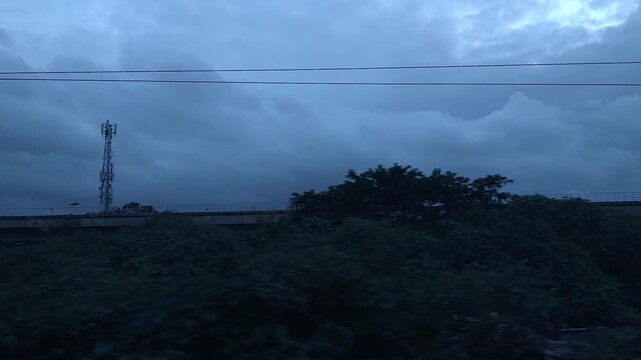Dramatic stormy skies loom over an urban industrial landscape with an elevated bridge and factories, captured from a moving perspective in India.