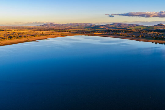 Aerial view showcasing the expansive waters of Lake Keepit, leading towards the surrounding rural landscape illuminated by soft morning light in NSW, Australia.