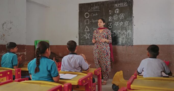 Indian young adult female talk speak teach to girl boy group class room board indoor rural area teen child wear uniform sitting on bench look tutor follow learn poem action moves enjoy day desi life