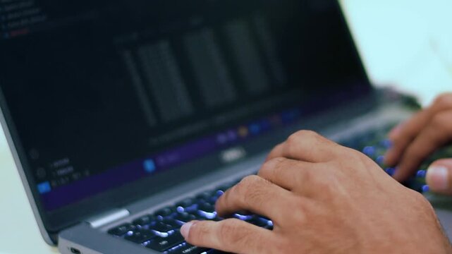 Close-up of a person's hands rapidly typing on a laptop keyboard with glowing keys, working on code or data in a modern office environment.