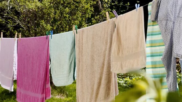 Colorful towels dry on a backyard clothesline in warm sunlight, clipped with pegs above green grass beside bushes and garden shade.