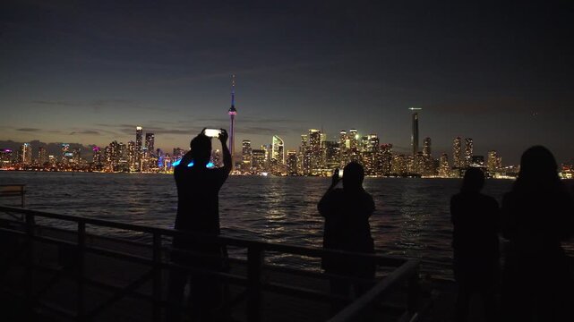 Silhouettes of tourists photographing the Toronto skyline from the waterfront as the camera slowly moves forward on a gimbal at dusk.