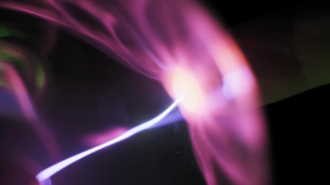 Macro view of a finger touching a glass globe and attracting a bright white electric arc. Pink plasma tendrils follow the contact point demonstrating high voltage conductivity.