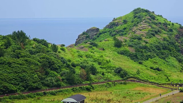 Elevated close-up over lush green hill with layered volcanic rock strata and wooden boardwalk trail by blue sea at Yongmeori Beach in Jeju, South Korea