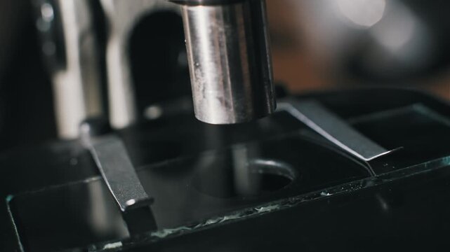 Macro shot of fingers placing a glass slide on a microscope stage secured by metal clips. A pipette adds a clear liquid for chemical analysis or a scientific procedure.