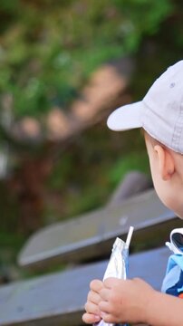 Two-year-old toddler in cap sits on the bench outdoors. Kid holds a doy pack and drinks from it with a straw. Side view. Vertical video