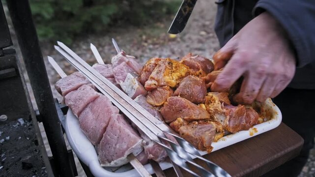 High-angle close-up of male hands mixing raw pork chunks with spices in a white tray. The meat is coated in orange seasoning representing preparation for a traditional outdoor barbecue.