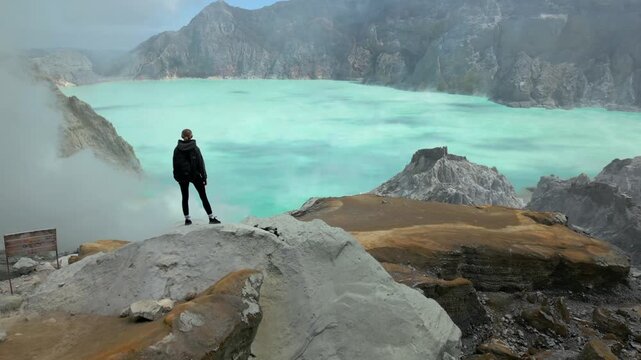 Young woman hiking along the rim of Kawah Ijen crater overlooking the turquoise acidic lake in East Java, Indonesia. Dramatic volcanic landscape with rugged cliffs and sulfur mist.