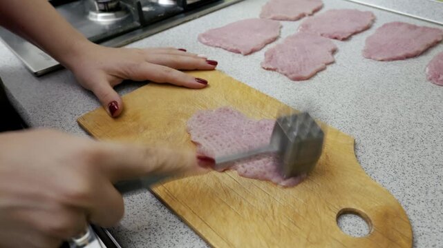Female hands use a metal meat mallet to tenderize a raw pork cutlet on a wooden board in a kitchen. Several other flattened pieces of meat are arranged on the countertop in the background.